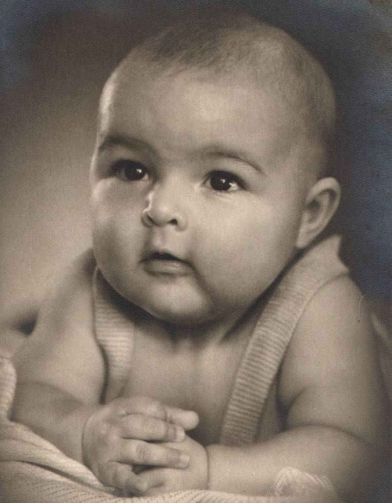 Schwarz/Weiß-Foto. Ein Baby aufgestützt auf seine Arme schaut neugierig drein.
